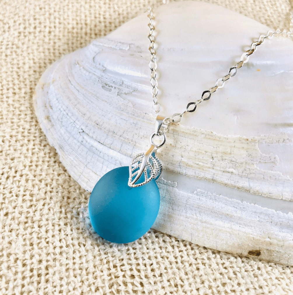 A turquoise sea glass pendant with a silver leaf design, hanging on a delicate silver chain, placed on a textured natural shell and a burlap background.